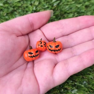 Può includere: Tre zucche in miniatura di colore arancione con volti di jack-o'-lantern intagliati, caratterizzati da occhi neri e bocche frastagliate. Ogni zucca ha un piccolo gambo marrone sulla parte superiore. Questi piccoli oggetti decorativi sono lisci.