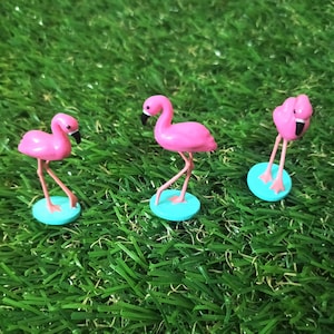 May include: Three pink flamingo ornaments with black beaks and long, thin legs. Each flamingo stands on a small, round, light blue base. The ornaments are set against a backdrop of green artificial grass.