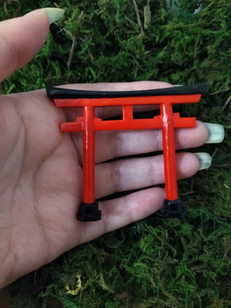 May include: A miniature red and black torii gate, a traditional Japanese gateway, is held in a hand. The gate is made of wood and has a black top and red pillars.