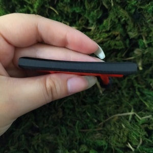 May include: A black and red plastic key chain with a small, detailed design. The keychain is held in a hand.