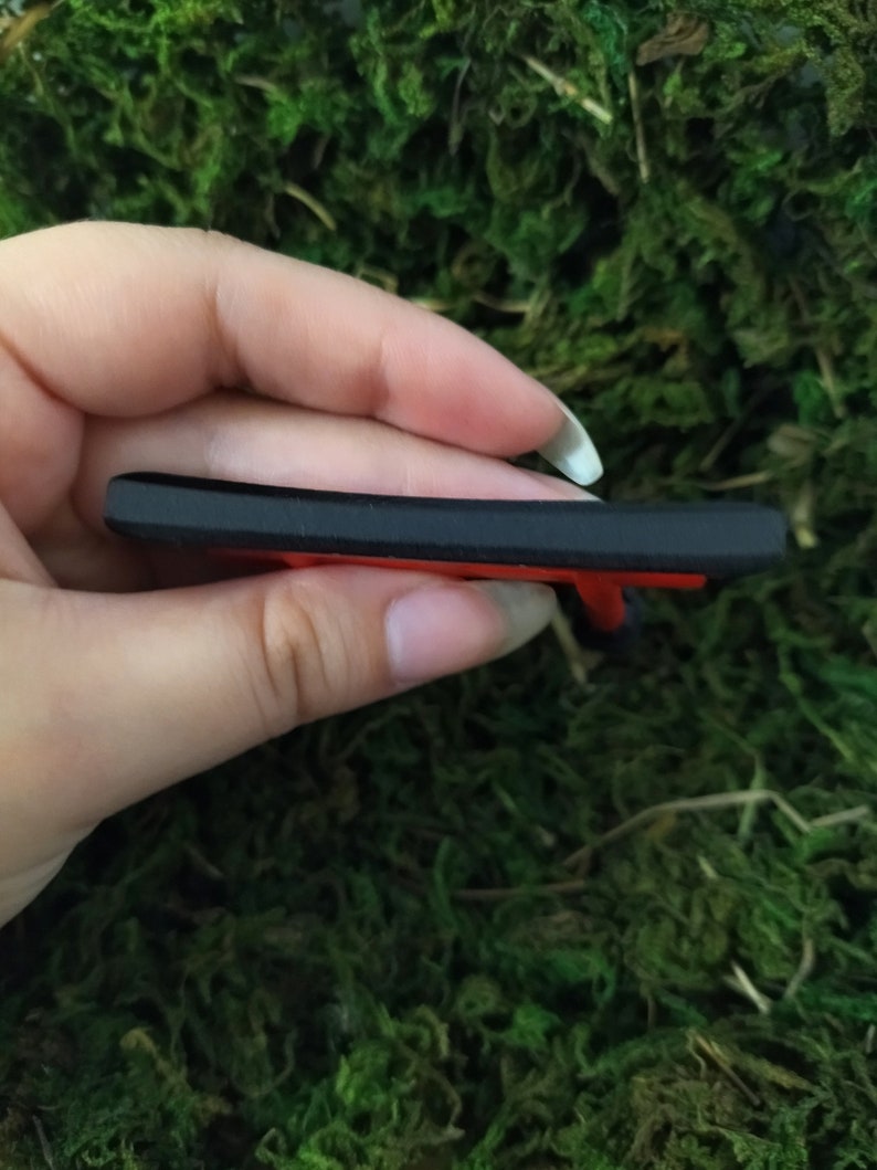 May include: A black and red plastic key chain with a small, detailed design. The keychain is held in a hand.