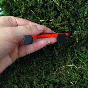 May include: A red and black miniature skateboard with black wheels. The skateboard is being held in a hand.