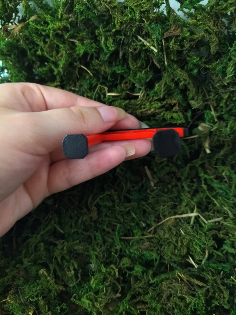 May include: A red and black miniature skateboard with black wheels. The skateboard is being held in a hand.