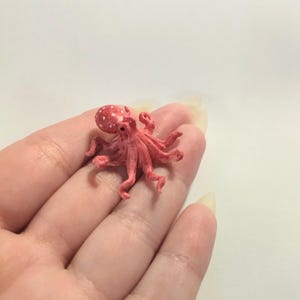 Peut inclure: Une petite figurine d'une pieuvre rouge avec des points blancs. La pieuvre a huit tentacules et est posée sur une main.