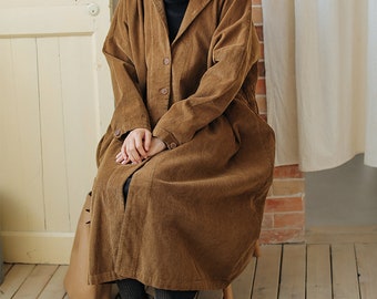 Può includere: Un lungo cappotto di velluto a coste marrone con chiusura a bottoni e colletto. Il cappotto è indossato da una persona seduta su una sedia.