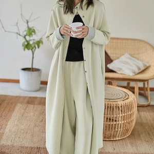 May include: A long, light green button-up shirt with a collar and long sleeves. The shirt has a relaxed fit and is made of a soft, comfortable fabric. The shirt is shown being worn by a person holding a white cup.