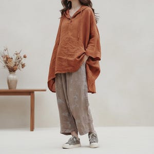 Op de afbeelding: Een roestkleurige hoodie met een relaxte pasvorm, gecombineerd met een loszittende, lichtbruine broek. De outfit wordt gecompleteerd met grijze sneakers. De kleding lijkt gemaakt van linnen of een vergelijkbare lichtgewicht stof, geschikt voor vrijetijdskleding.