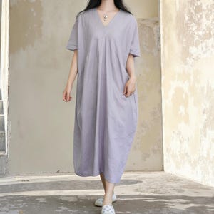 May include: A lilac-coloured, short-sleeved dress with a V-neck. The dress has a loose, flowing design with pockets and falls to the mid-calf. The model is wearing white shoes. The dress is made of a soft, lightweight fabric.