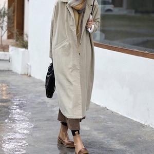May include: A long, beige corduroy coat with a single button closure and a patch pocket on the left side. The coat is worn over a brown and white patterned skirt and brown ankle boots.