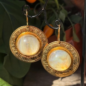 Pode incluir: Brincos dourados com uma pedra redonda iridescente engastada em uma moldura circular com um design ornamentado.