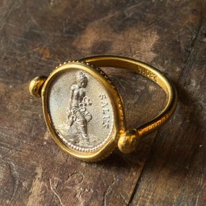 Può includere: Anello d'oro con una faccia ovale con un design dettagliato di moneta d'argento. La moneta raffigura una figura con testo. La fascia dell'anello è liscia e arrotondata, con piccole sfere decorative.