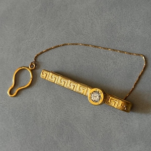 Op de afbeelding: Een goudkleurige stropdasklem met een geometrisch patroon en een centrale, ronde, heldere edelsteen. De klem is via een fijne ketting verbonden met een kleine lus, ontworpen om een stropdas aan een overhemd te bevestigen.