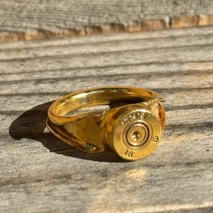 May include: A gold ring with a bullet casing design. The bullet casing has the number 18 on it.