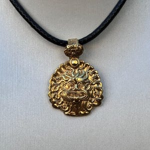 Könnte beinhalten: Eine goldene Halskette mit einem runden Anhänger, der ein detailliertes, geprägtes Design eines mythischen Gesichts zeigt. Der Anhänger ist an einem schwarzen Lederband befestigt.