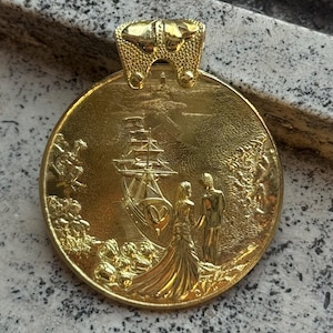 Może przedstawiać: Złoty wisiorek z szczegółową sceną reliefową przedstawiającą statek, parę i liście. Wisiorek ma teksturowaną krawędź u góry do zawieszenia. Projekt jest skomplikowany, z naciskiem na żagle statku i strój pary.