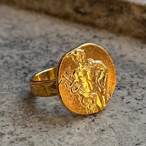 Könnte beinhalten: Ein goldfarbener Ring mit einer großen, runden Oberfläche, die ein detailliertes Relief einer klassischen Figur mit Stab und Schlange zeigt. Der Ring hat ein strukturiertes Design. Der Ring befindet sich vor einem neutralen Hintergrund.