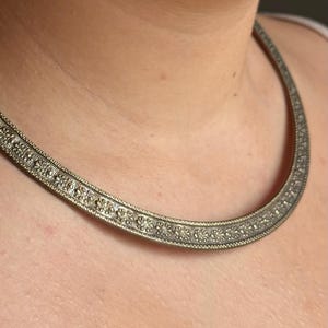 Può includere: Una collana color argento con un motivo floreale. La collana è uno stile girocollo, che poggia vicino al collo. Il metallo ha un aspetto leggermente invecchiato, con dettagli intricati. Il design presenta piccoli motivi floreali ripetuti.
