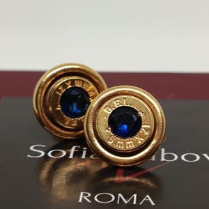 Könnte beinhalten: Ein Paar goldplattierte Kugelohrringe mit blauen Edelsteinen. Die Ohrringe sind rund und haben einen erhöhten Rand. Die Patronenhülsen sind mit "9mm" und "12 gauge" gekennzeichnet.