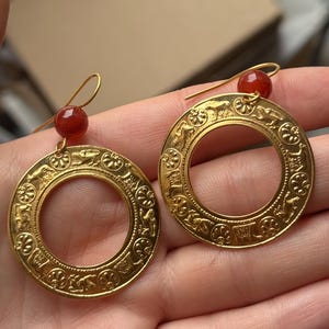 Peut inclure: Paire de boucles d'oreilles dorées en forme de cerceau, ornées de motifs en relief complexes et d'une petite perle rouge. Les boucles d'oreilles ont une forme circulaire et une fermeture à crochet.