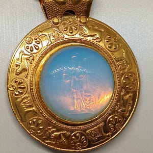 Op de afbeelding: Een gouden ketting met een ronde, blauwe glazen cameo met een vrouw die een staf en schild vasthoudt. De hanger is omgeven door een decoratieve rand met dierlijke en bloemmotieven.