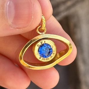 May include: A gold-coloured eye-shaped pendant with a blue gemstone centre. The gemstone is set within a gold-coloured casing, with the text "GFI 9mm" visible. The pendant is suspended from a small loop.