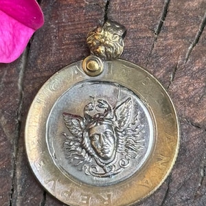 Può includere: Ciondolo in stile vintage con una moneta d'argento dettagliata raffigurante un volto alato. La moneta è incastonata in una cornice dorata e sormontata da una piccola e ornata testa di leone. Un gioiello unico.