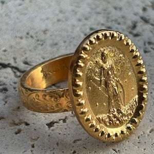 Op de afbeelding: Een goudkleurige ring met een ovale voorkant met een gedetailleerd reliëf van een figuur. De ringband heeft een gegraveerd bloemmotief en het woord "ROMA" is zichtbaar. De ring ligt op een getextureerd, lichtgekleurd oppervlak.