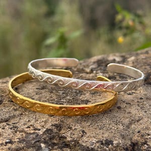 Può includere: Due braccialetti, uno dorato e uno argentato, con un design a onde. I braccialetti sono impilati l'uno sull'altro.