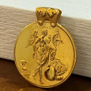 Könnte beinhalten: Ein goldfarbener Anhänger mit einem gravierten Design. Der Anhänger zeigt eine geflügelte Figur, die ein Rad und einen Globus hält, mit dem Wort "FORTUNA" oben. Ein Schiff und ein Gebäude sind ebenfalls abgebildet.