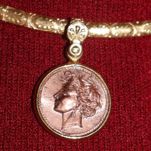 Può includere: Una collana dorata con un pendente circolare raffigurante il profilo di una donna. Il pendente è color rame con un bordo dorato. La collana ha una fascia decorativa strutturata e un accento a giglio.