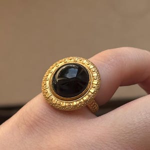 May include: A gold ring with a large, round, black gemstone. The ring has a textured band and a decorative bezel. The ring is shown on a finger. The ring's design is ornate.