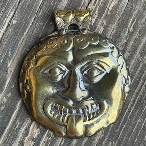 May include: A gold-toned metal pendant featuring a detailed, stylized face with a wide, toothy grin and a protruding tongue. The pendant has a decorative, ornate top.