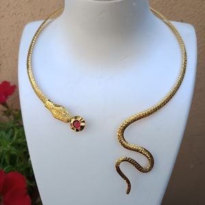 Op de afbeelding: Een gouden slangen choker ketting met een rode edelsteen accent. De slang is opgerold en heeft een getextureerd oppervlak.