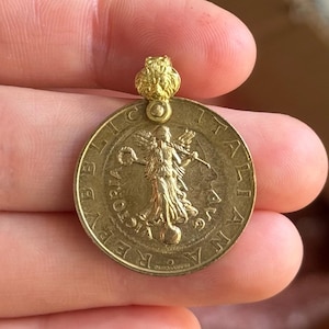 Op de afbeelding: Goudkleurige hanger met een munt met een afbeelding van een gevleugelde figuur en de woorden "REPUBBLICA ITALIANA". De hanger heeft een kleine leeuwenkop aan de bovenkant.