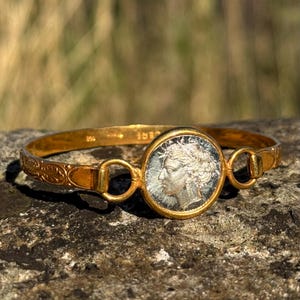 Può includere: Bracciale dorato con una moneta d'argento raffigurante un ritratto di profilo. Il bracciale presenta un design dettagliato con elementi decorativi e un'incastonatura circolare per la moneta. I dettagli della moneta sono visibili.