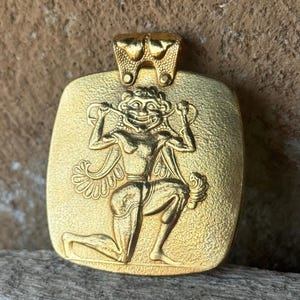 Può includere: Ciondolo color oro con superficie strutturata. Il ciondolo presenta un rilievo dettagliato di una figura inginocchiata con volto sorridente e dettagli ornati. Il passante è di design decorativo. Il ciondolo è probabilmente in metallo.