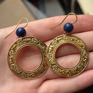 Peut inclure: Une paire de boucles d'oreilles dorées avec des motifs en relief complexes et une petite perle bleu foncé au sommet. Les boucles d'oreilles ont une fermeture à crochet et sont présentées sur un fond neutre.