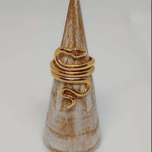 Könnte beinhalten: Ein goldfarbener Schlangenring mit strukturierter Oberfläche. Der Ring ist um einen hölzernen Ringständer gewickelt.