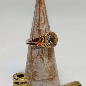 Può includere: Un anello d'oro realizzato con un bossolo di proiettile .38 Special con una pietra preziosa trasparente incastonata al centro. L'anello è esposto su un supporto per anelli in legno. Due ulteriori bossoli di proiettile sono visibili in primo piano.
