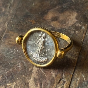 Op de afbeelding: Een gouden ring met een cirkelvormige face met een zilveren figuur en sterren. De ring heeft een dikke band en twee gouden bollen aan weerszijden van de face. De ring ligt op een houten oppervlak.