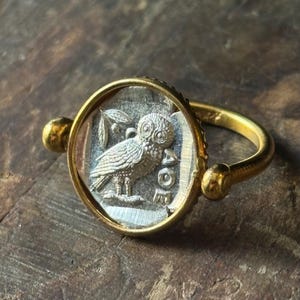 Puede incluir: Anillo dorado con un diseño detallado de búho plateado dentro de un engaste claro y circular. El búho, con detalles de plumas, sostiene una ramita. El anillo tiene una banda pulida y elementos decorativos. El diámetro es de unos 2 cm.