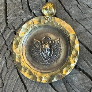 Può includere: Un ciondolo rotondo color oro con una superficie testurizzata. Il ciondolo presenta un'immagine in rilievo di una figura femminile alata con un serpente nei capelli. Il viso della figura è visibile e ha un'espressione seria.