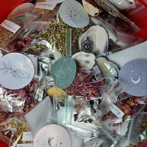 Puede incluir: Un bol de plástico rojo lleno de pequeñas bolsas de plástico transparentes que contienen diversas hierbas secas, flores y cristales. Las bolsas están etiquetadas con texto escrito a mano, incluyendo "rosewood" y "moonflower". También hay varias pegatinas blancas pequeñas con ilustraciones dibujadas a mano de manos, lunas y estrellas.