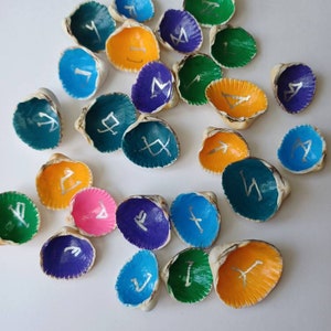 Puede incluir: Una colección de conchas marinas pintadas, cada una con un interior de diferente color y un símbolo rúnico plateado. Los colores incluyen azul, verde, naranja, morado y rosa. Las conchas están dispersas sobre una superficie blanca.