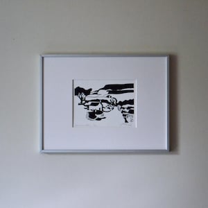 May include: A framed black and white landscape print. The artwork depicts a stylized scene with trees, a building, and rolling hills. The print is matted and set in a silver frame, mounted on a white wall.