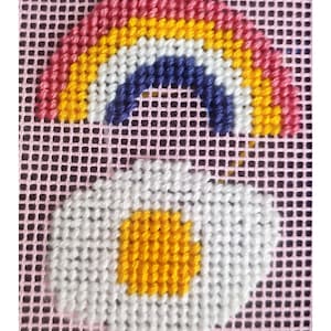 Peut inclure: Motif de point de croix repr&eacute;sentant un arc-en-ciel et une marguerite. L'arc-en-ciel est en haut, dans des tons de rose, jaune, bleu et blanc. En dessous, une marguerite blanche avec un centre jaune est affich&eacute;e sur un fond rose.