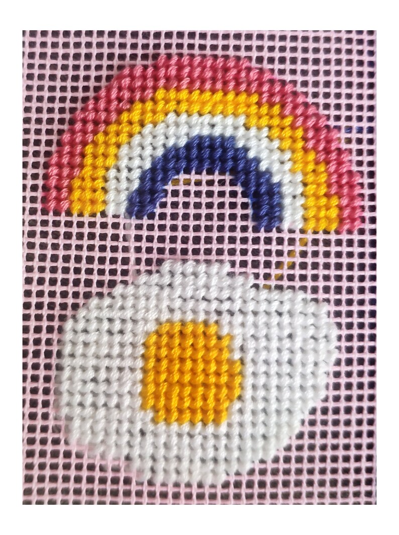 Peut inclure: Motif de point de croix repr&eacute;sentant un arc-en-ciel et une marguerite. L'arc-en-ciel est en haut, dans des tons de rose, jaune, bleu et blanc. En dessous, une marguerite blanche avec un centre jaune est affich&eacute;e sur un fond rose.