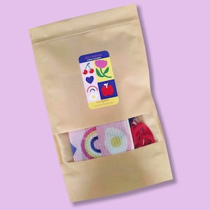 Peut inclure: Un kit de bricolage dans un sac en papier brun avec une fen&ecirc;tre transparente. Le kit comprend une toile avec un motif arc-en-ciel et du fil rouge. Une &eacute;tiquette sur le sac indique "KIT CANEVAS" et pr&eacute;sente des illustrations color&eacute;es.