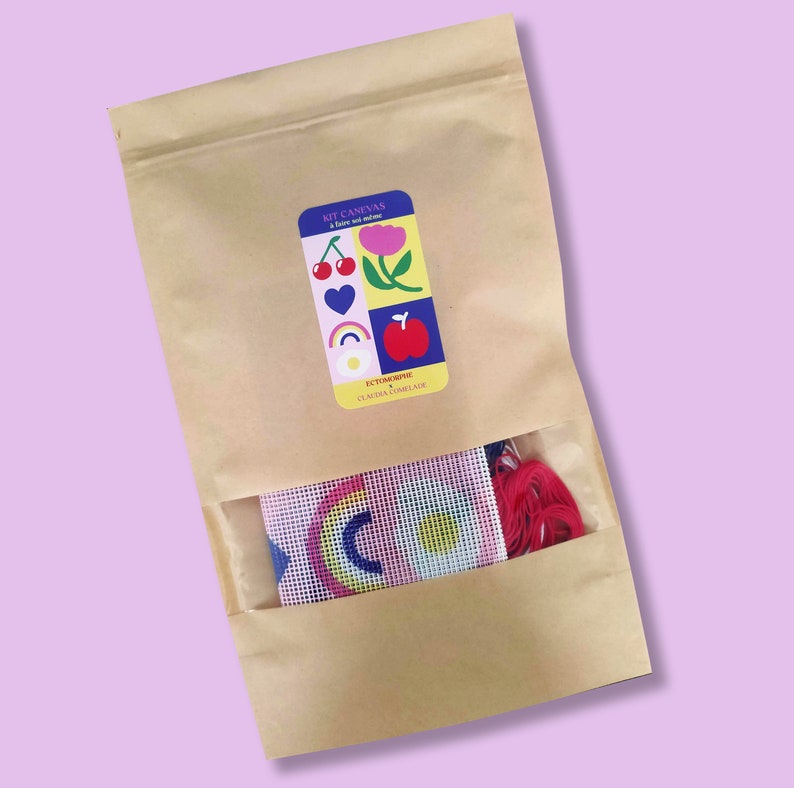 Peut inclure: Un kit de bricolage dans un sac en papier brun avec une fen&ecirc;tre transparente. Le kit comprend une toile avec un motif arc-en-ciel et du fil rouge. Une &eacute;tiquette sur le sac indique "KIT CANEVAS" et pr&eacute;sente des illustrations color&eacute;es.