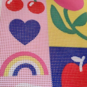 Peut inclure: Une toile de point de croix color&eacute;e avec un motif r&eacute;p&eacute;t&eacute; de cerises, un c&oelig;ur bleu, un arc-en-ciel, une fleur et une pomme rouge. Le fond est divis&eacute; en carr&eacute;s roses et jaunes, avec une texture quadrill&eacute;e.